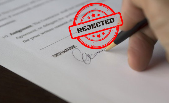 A hand signing a document with a stamp saying "rejected"
